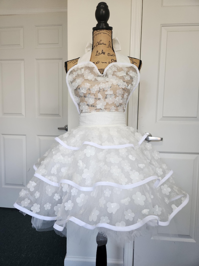 May include: A white apron featuring a halter neck design with a sweetheart neckline. The bodice and the three-tiered skirt are adorned with white floral appliqu&eacute;s and sheer mesh fabric, finished with solid white ribbon trim along the edges.