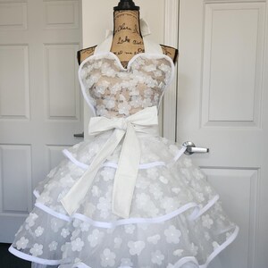 May include: A white apron featuring a halter neck design with a large waist bow. The apron is crafted from sheer fabric adorned with layered white floral appliqu&eacute;s and finished with solid white trim along the tiered, ruffled skirt edges.