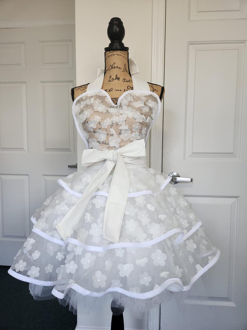 May include: A white apron featuring a halter neck design with a large waist bow. The apron is crafted from sheer fabric adorned with layered white floral appliqu&eacute;s and finished with solid white trim along the tiered, ruffled skirt edges.