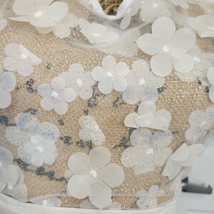 May include: A white garment featuring a sheer mesh overlay adorned with numerous three-dimensional white floral appliqu&eacute;s. The fabric has a subtle glittery finish throughout, and the neckline is trimmed with a solid white fabric border.