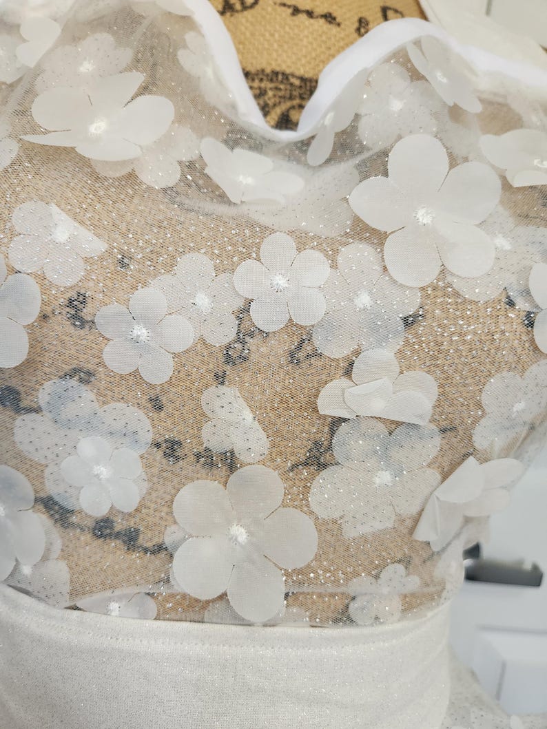 May include: A white garment featuring a sheer mesh overlay adorned with numerous three-dimensional white floral appliqu&eacute;s. The fabric has a subtle glittery finish throughout, and the neckline is trimmed with a solid white fabric border.