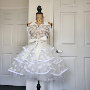 May include: A white apron featuring a sweetheart neckline, a large waist tie, and a multi-layered skirt. The fabric is adorned with small, white, dimensional floral appliqu&eacute;s and finished with solid white trim along the edges of the tiers.