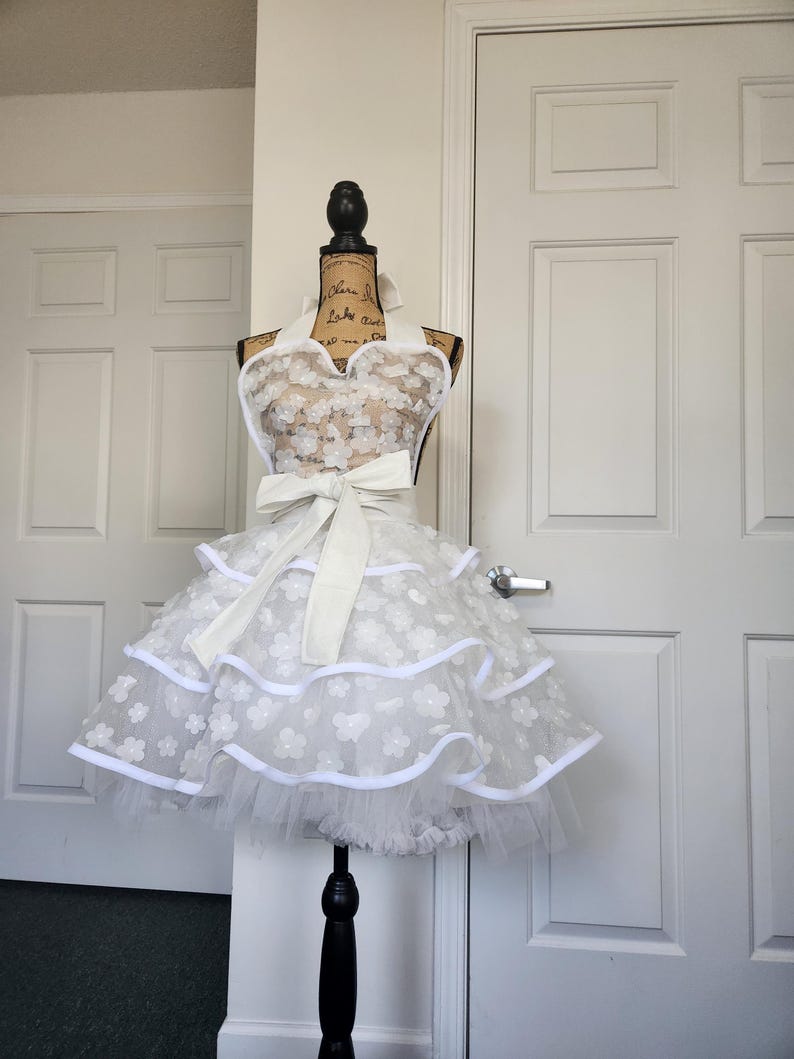 May include: A white apron featuring a sweetheart neckline, a large waist tie, and a multi-layered skirt. The fabric is adorned with small, white, dimensional floral appliqu&eacute;s and finished with solid white trim along the edges of the tiers.