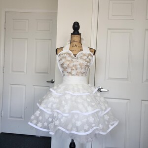 May include: A white apron featuring a sweetheart neckline and a tiered skirt. The bodice and skirt are adorned with layered white floral appliqu&eacute;s. The apron includes a wide white waistband and white ribbon trim along the edges of the tiers.