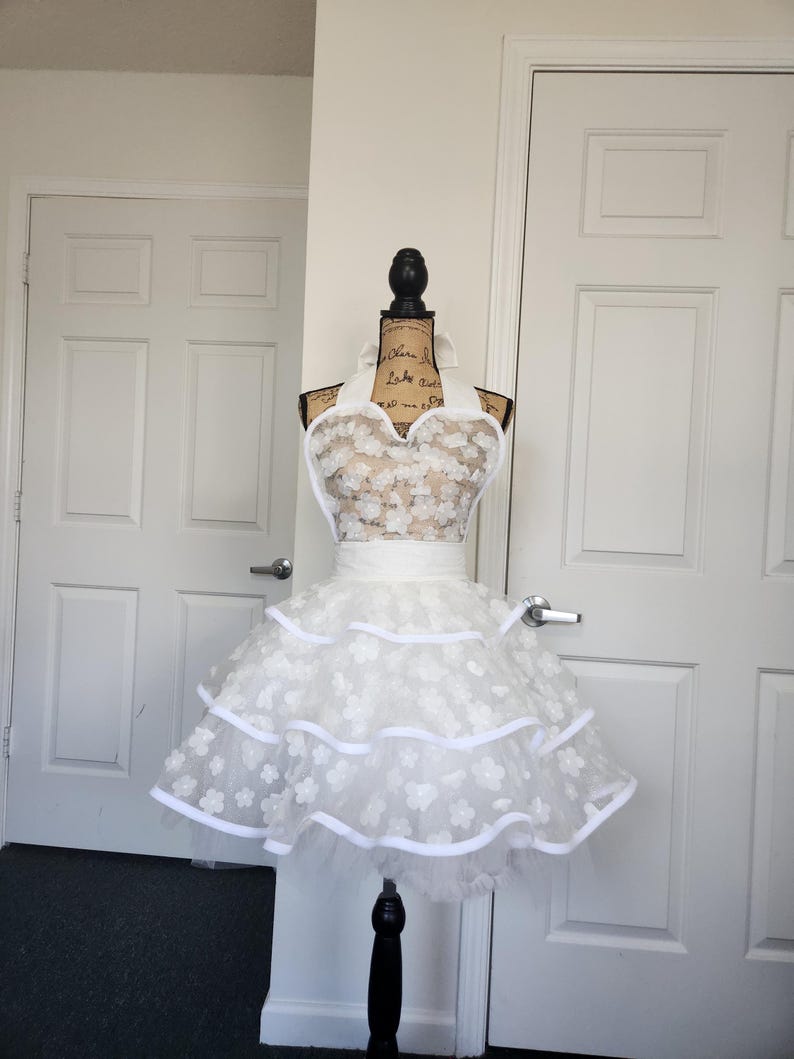May include: A white apron featuring a sweetheart neckline and a tiered skirt. The bodice and skirt are adorned with layered white floral appliqu&eacute;s. The apron includes a wide white waistband and white ribbon trim along the edges of the tiers.