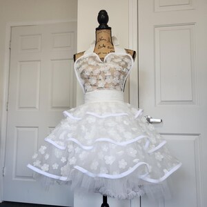 May include: A white apron featuring a halter neck design and a bodice covered in small floral appliqu&eacute;s. The skirt consists of three tiered layers of white tulle, each trimmed with a solid white ribbon edge, creating a full and voluminous silhouette.