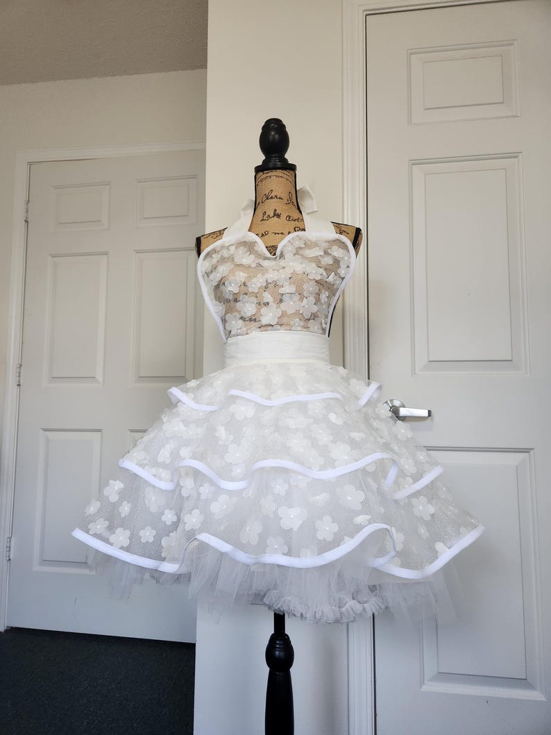 May include: A white apron featuring a halter neck design and a bodice covered in small floral appliqu&eacute;s. The skirt consists of three tiered layers of white tulle, each trimmed with a solid white ribbon edge, creating a full and voluminous silhouette.