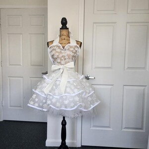 May include: A white apron featuring a sweetheart neckline, a large tied bow at the waist, and a multi-layered skirt. The fabric is adorned with a repeating pattern of small white floral appliqu&eacute;s and finished with solid white trim along the edges.