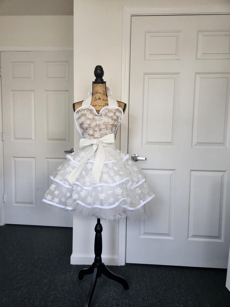 May include: A white apron featuring a sweetheart neckline, a large tied bow at the waist, and a multi-layered skirt. The fabric is adorned with a repeating pattern of small white floral appliqu&eacute;s and finished with solid white trim along the edges.