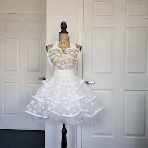 May include: A white apron featuring a halter neck design with a bodice and a two-tiered skirt covered in small, dimensional floral appliqu&eacute;s. The apron includes a solid white waistband and white ribbon trim along the edges of the tiers.