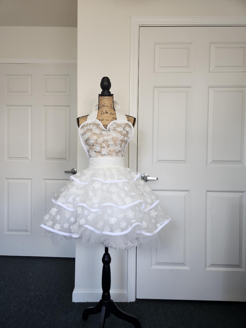 May include: A white apron featuring a halter neck design with a bodice and a two-tiered skirt covered in small, dimensional floral appliqu&eacute;s. The apron includes a solid white waistband and white ribbon trim along the edges of the tiers.