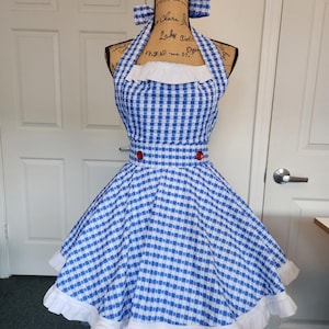 May include: A blue and white gingham apron with a halter neck and a flared skirt. The apron features a white ruffle trim along the bottom and red buttons at the waist. Two small bows are attached to the top of the apron.