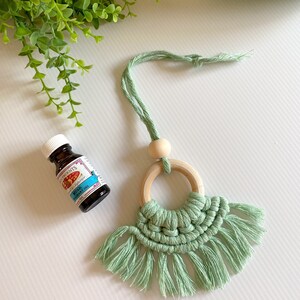 May include: A light green macrame car air freshener with a wooden ring and bead. A small bottle of essential oil is next to the air freshener. The oil bottle has a white cap and a blue label with text.