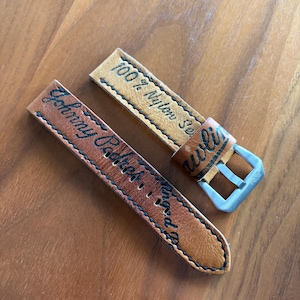May include: Two brown leather watch straps with black stitching and text. One strap reads "Johnny Cakes & Co." and the other reads "100% Nylon Sewn". A silver-colored buckle is attached to one strap.