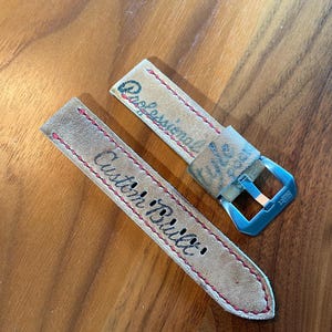 May include: A pair of tan leather watch straps with red stitching. One strap reads "Professional" and the other reads "Custom Built" in black script. A blue metal buckle is attached to one strap.