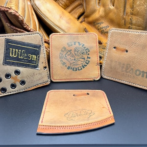 May include: A collection of vintage baseball glove leather patches and accessories. Includes patches with the "Wilson" logo, "Style Pocket" graphic, and other details. The items are in various shapes and sizes, with visible stitching and wear.