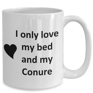 May include: White ceramic coffee mug with a black heart and the text "I only love my bed and my Conure" printed on it.