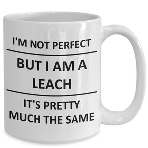 May include: White ceramic mug with a curved handle. The mug features black text that reads "I'M NOT PERFECT BUT I AM A LEACH IT'S PRETTY MUCH THE SAME". The mug is designed for hot beverages.