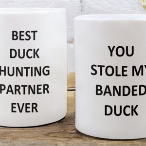 May include: Two white ceramic mugs with black text. One mug reads "BEST DUCK HUNTING PARTNER EVER." The other mug says "YOU STOLE MY BANDED DUCK."