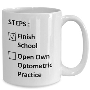 May include: White ceramic coffee mug with black text that reads "STEPS: Finish School Open Own Optometric Practice". A tick is next to "Finish School" and a square is next to "Open Own Optometric Practice".
