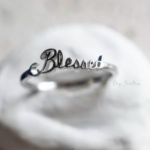 Könnte beinhalten: Ein silberner Ring mit dem Wort "Blessed" in Schreibschrift. Der Ring wird auf einer weißen, strukturierten Oberfläche präsentiert. Das Design des Rings ist schlicht und elegant, geeignet für den Alltag.