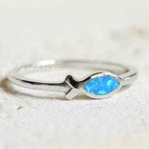 May include: A silver ring featuring a fish-shaped design with a vibrant blue opal inlay. The ring is set against a neutral background, highlighting the delicate details of the jewelry. The text "6 Day Creations" is visible in the background.