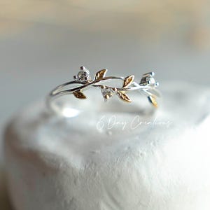 Puede incluir: Un anillo de plata con un delicado diseño de hojas y vid. El anillo presenta pequeñas piedras preciosas transparentes y detalles de hojas doradas. El anillo se muestra sobre una superficie blanca texturizada. Es una joya.