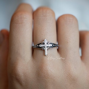 May include: A silver ring featuring a cross design, adorned with small, sparkling stones. The cross is the focal point, set against a detailed band with decorative elements. The ring is on a finger, showcasing the intricate craftsmanship.