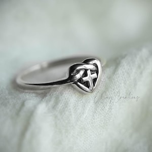Könnte beinhalten: Ein silberner Ring mit einem herzförmigen Design und einem Kreuz in der Mitte. Der Ring hat einen zarten Ring und eine polierte Oberfläche. Der Ring liegt auf einem weichen, strukturierten Hintergrund.