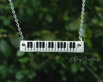 Puede incluir: Un collar de plata con un colgante de teclas de piano. El colgante es una barra de plata con teclas de piano negras.