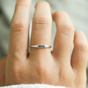 Puede incluir: Un anillo plateado con un pequeño diseño rectangular en la parte superior. El anillo se lleva en un dedo y el fondo está borroso. El anillo parece estar hecho de un metal brillante.