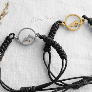 Könnte beinhalten: Zwei schwarze Kordelarmbänder mit einem Berg-Silhouetten-Anhänger. Ein Anhänger ist silber mit einer goldenen Sonne, der andere ist gold mit einer goldenen Sonne.