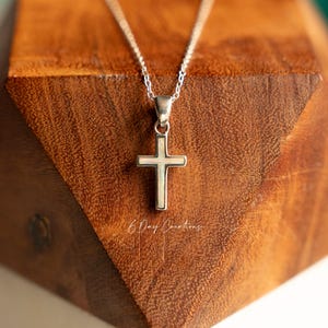 May include: A silver cross pendant necklace with a delicate chain. The cross has a pearlescent center and is displayed on a wooden block. The text "6 Day Creations" is visible on the wood.