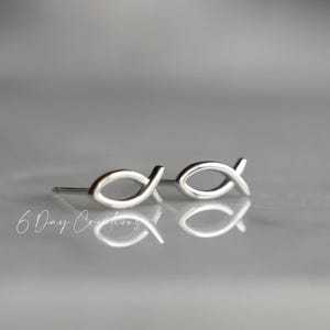 May include: A pair of silver-colored stud earrings in the shape of the Christian fish symbol. The earrings are displayed on a reflective surface. The text "6 Day Creations" is visible in the lower left corner.
