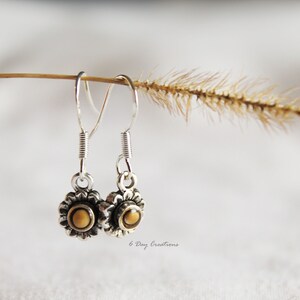 May include: Silver dangle earrings with a small sunflower design featuring a brown centre. The earrings are hanging from a twig with a brown seed head.