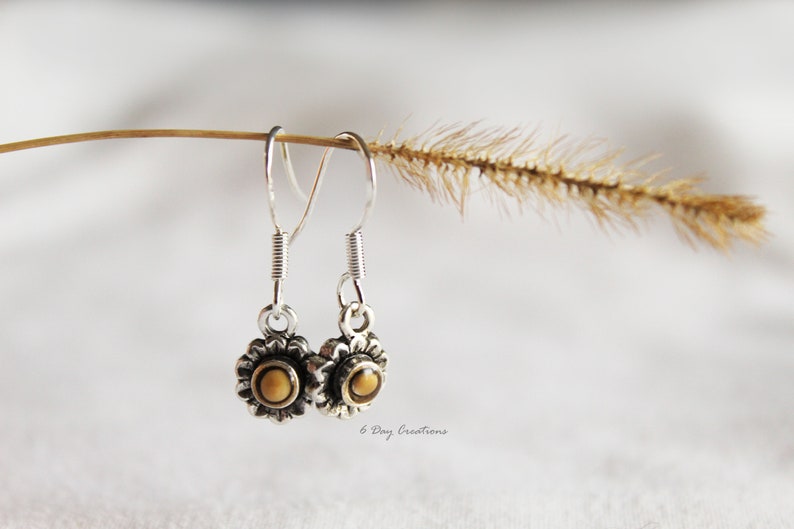 May include: Silver dangle earrings with a small sunflower design featuring a brown centre. The earrings are hanging from a twig with a brown seed head.