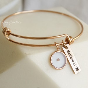 Puede incluir: Pulsera de brazalete de oro con un pequeño dije redondo y un dije rectangular con el texto "Mateo 17:20".