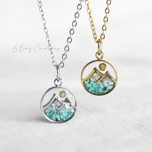 May include: Two necklaces with circular pendants. One is silver, the other gold. Each pendant features a mountain scene with turquoise and white stones, and a yellow sun. The necklaces are on a white background.