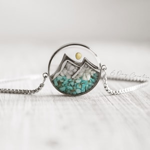 May include: A silver chain bracelet with a round pendant featuring a mountain scene with turquoise and white stones. The pendant also has a small yellow sun.