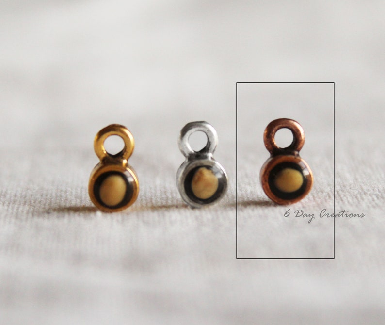 Pu&ograve; includere: Tre piccoli ciondoli in metallo con un centro rotondo di colore marrone chiaro e un bordo nero. I ciondoli sono in oro, argento e rame.