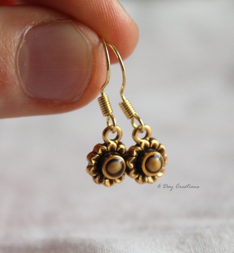 May include: Gold dangle earrings with a small flower design and a brown centre. The earrings have a spring-loaded hook closure.