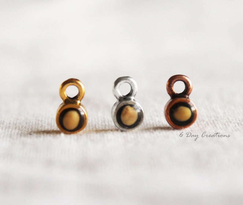 Pu&ograve; includere: Tre piccoli ciondoli con un centro rotondo color crema e un bordo nero. I ciondoli sono attaccati a un anello e sono disponibili in oro, argento e rame.