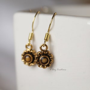 May include: A pair of gold dangle earrings with a small, round, brown and gold flower design. The earrings have a spring closure.