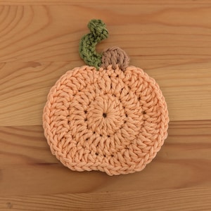 May include: A handmade, crocheted pumpkin coaster in a light orange colour. The pumpkin has a green stem and a brown top. The coaster is circular and sits on a wooden surface.