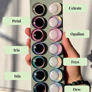 May include: A selection of colorful, round craft eyes in various shades, including black, pink, green, and blue, are displayed on a green card. Each eye has a black pupil and a shimmering, iridescent iris. The names "Celeste", "Petal", "Opaline", "Iris", "Feya", "Isla", and "Dew" are labeled.