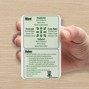 May include: A green and white card with a disc golf disc in the center and arrows pointing in different directions. The card has the words "Wind", "Headwind", "Tailwind", "From Left", "From Right", and "Notes". The text on the card explains how wind affects disc golf throws.