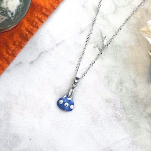 Puede incluir: Un collar plateado con un colgante azul con lunares blancos. El colgante tiene forma triangular pequeña. El collar se exhibe sobre una superficie de mármol blanco con tela naranja y plantas secas en el fondo.