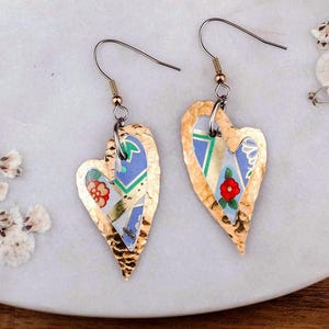 Pode incluir: Um par de brincos pendentes em forma de coração com uma borda de metal dourado martelado envolvendo uma incrustação central de material com padrão azul, branco e verde com pequenos detalhes florais vermelhos e laranjas, suspensos por ganchos finos.