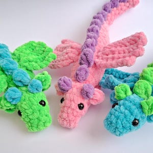 Peut inclure: Trois dragons en crochet en vert, rose et bleu. Chaque dragon a un corps textur&eacute;, des ailes et de petites oreilles. Les dragons sont pos&eacute;s sur une surface blanche. Les dragons sont faits d'une mati&egrave;re douce et moelleuse.