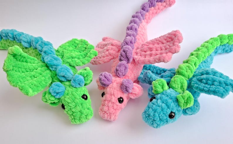 Peut inclure: Trois dragons en crochet en vert, rose et bleu. Chaque dragon a un corps textur&eacute;, des ailes et de petites oreilles. Les dragons sont pos&eacute;s sur une surface blanche. Les dragons sont faits d'une mati&egrave;re douce et moelleuse.